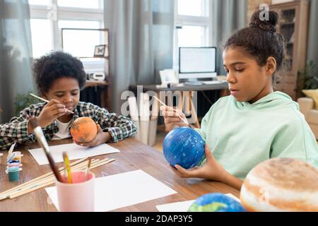 Two adorable mixed-race siblings in casualwear painting planets by table at home Stock Photo