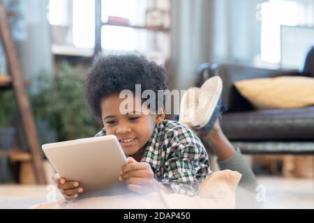 Happy elementary schoolboy with tablet lying on the floor by black leather couch Stock Photo