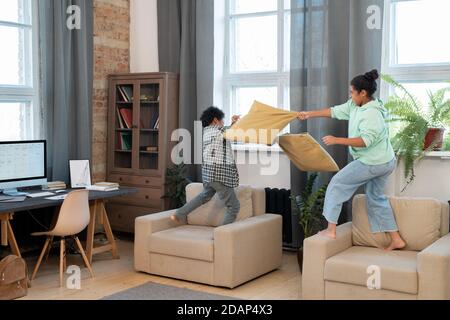 Two cute mixed-race siblings in casualwear pillow fighting on soft armchairs Stock Photo