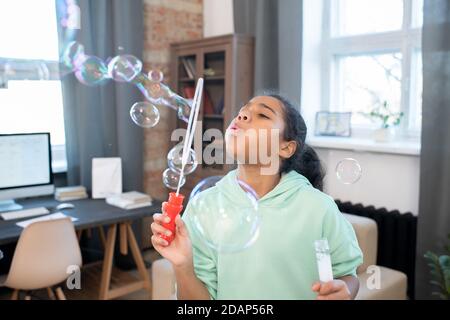 Cute mixed-race girl in casualwear blowing group of soap bubbles in living-room Stock Photo