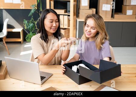 Happy young Asian woman taking photo of headphones with her colleague near by Stock Photo