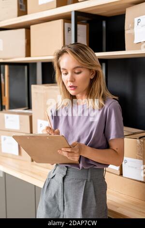 Young serious blond female with pen and clipboard checking orders of clients Stock Photo