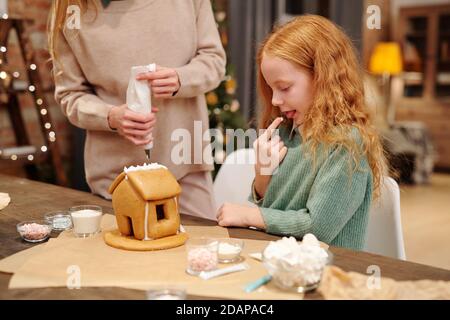 Adorable little girl tasting whipped cream from homemade gingerbread house Stock Photo