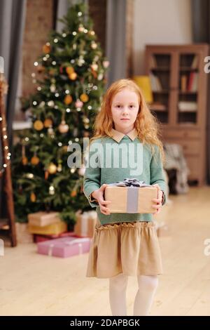 Adorable girl in casualwear holding giftbox with Christmas present from Santa Stock Photo