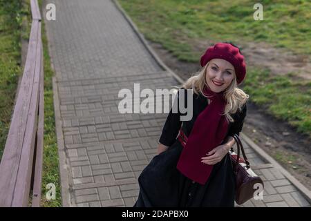 A joyful woman walks up the stairs in burgundy palla and biret, an adult with a beautiful smile in black clothes, in the fall against a blue sky. Stock Photo