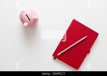 Creative flat lay photo of red notebook, pen and piggy bank on a white minimalistic background with copy space. Minimal work space Stock Photo