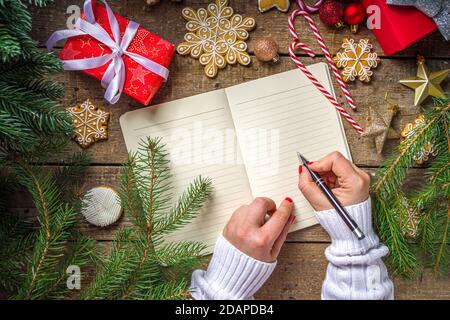 Woman hands write Christmas letter, wishlist or To Do list on notepad paper on wooden background with Christmas decoration, top view flatlay copy spac Stock Photo