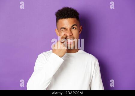 Image of young african-american man picking nose and looking upper left corner spaced out, standing over purple background Stock Photo