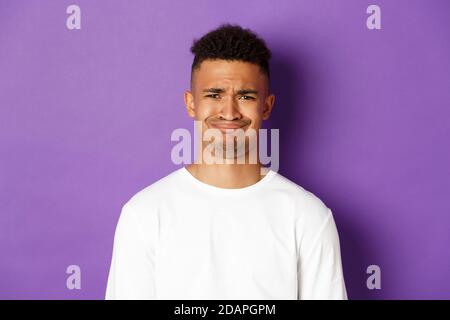 Close-up of skeptical and disappointed african-american man, grimacing and frowning dissatisfied, standing over purple background Stock Photo