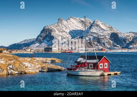 Red wooden cabin on raft with sailing boat in the sea off the coast, behind fishing village and mountains, Seloy, Nordland, Norway Stock Photo