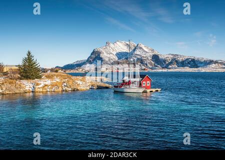 Red wooden cabin on raft with sailing boat in the sea off the coast, behind fishing village and mountains, Seloy, Nordland, Norway Stock Photo