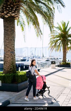 Mother is pushing a pink stroller with her baby inside in a marina in Montenegro Stock Photo