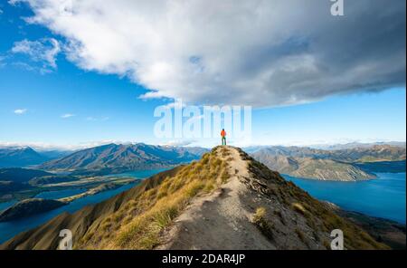 Hiker stands at the summit, view of mountains and lake from Mount Roy, Roys Peak, Lake Wanaka, Southern Alps, Otago, South Island, New Zealand Stock Photo