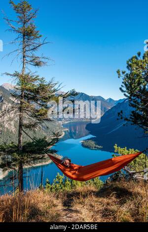 Young man lying in red hammock with panoramic view of mountains and lake, Plansee, Ammergau Alps, Reutte district, Tyrol, Austria Stock Photo