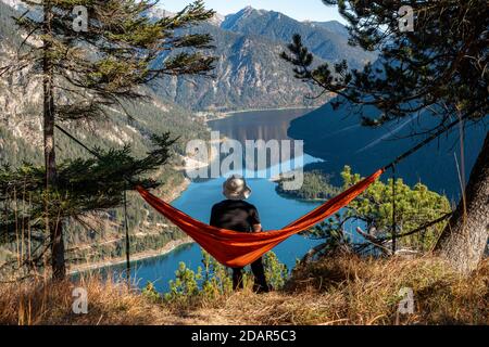 Young man sitting in red hammock with panoramic view of mountains and lake, Plansee, Ammergau Alps, Reutte district, Tyrol, Austria Stock Photo