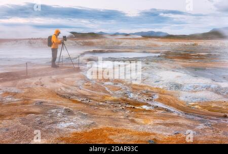 Man in yellow jacket with photo tripod, photo backpack and camera standing in the steaming red high temperature area Hveraroend with solfatars and Stock Photo