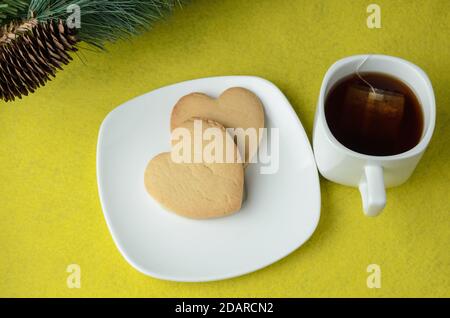Two heart-shaped cookies on a white plate with a mug of tea on a yellow background with a branch of an artificial Christmas tree with a cone. Selectiv Stock Photo