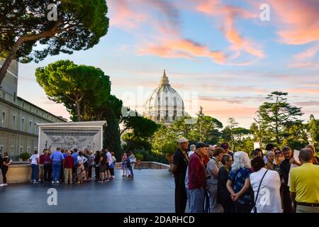 View of the dome of St Peters Basilica from the Vatican museum rooftop with groups of tourists and Italian stone pine trees under a colorful sky Stock Photo