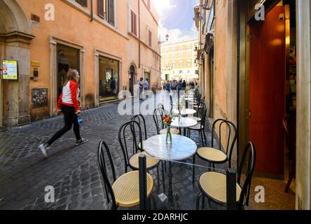 Tourists walk down a narrow cobbled street lined with patio outdoor tables from a cafe in the center of Rome, Italy Stock Photo