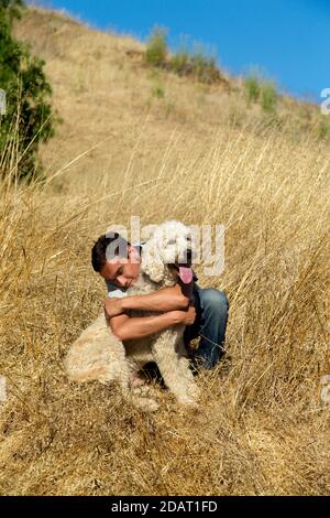 Young man with eyes closed hugging hi adult light colored Labradoodle on a wheat colored grass hill Stock Photo