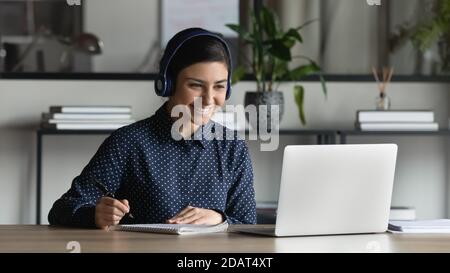 Smiling Indian businesswoman intern wearing headphones watching webinar, writing notes Stock Photo