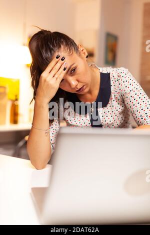 Businesswoman looking exhausted and massaging forhead while working overtime on laptop from home kitchen. Employee using modern technology at midnight doing overtime for job, business, career, network, lifestyle Stock Photo