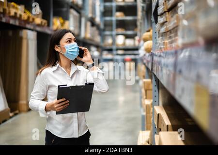 woman worker with medical mask talking on a mobile phone and holding clipboard to checking inventory in the warehouse during coronavirus (covid-19) pa Stock Photo