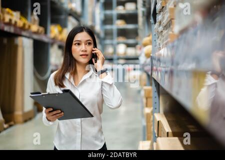 young woman worker talking on a mobile phone and holding clipboard to checking inventory in the warehouse store Stock Photo