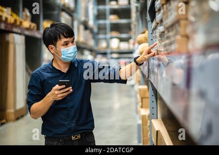 man worker with medical mask using smartphone to checking inventory in the warehouse during coronavirus (covid-19) pandemic Stock Photo