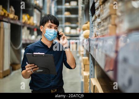man worker with medical mask talking on a mobile phone and holding clipboard to checking inventory in the warehouse during coronavirus (covid-19) pand Stock Photo