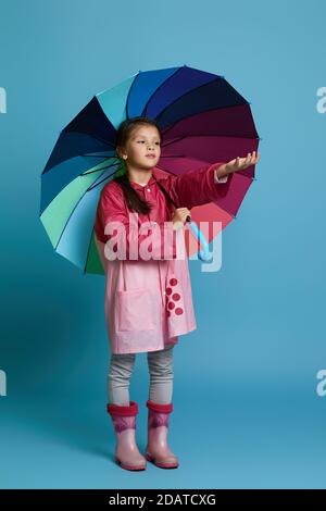 little child girl with multicolored umbrella in pink rain coat and rubber boots on blue background. girl checking for rain Stock Photo