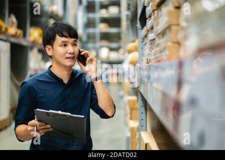 young man worker talking on a mobile phone and holding clipboard to checking inventory in the warehouse store Stock Photo