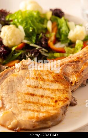 Fried pork entrecote on the bone with stewed pumpkin pieces and salad in a plate close up, green salad with cauliflower and napa cabbage, bell pepper Stock Photo