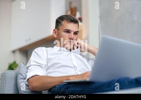 thoughtful young man browsing the Internet while lying on the couch. Stock Photo