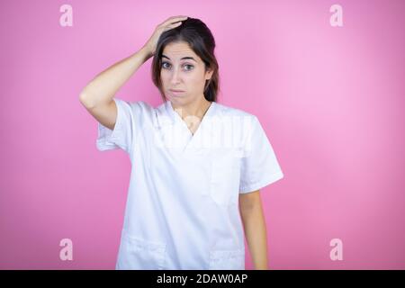 Young brunette doctor girl wearing nurse or surgeon uniform over isolated pink background putting one hand on her head smiling like she had forgotten Stock Photo