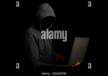 Man without a face in a hood holds a laptop on a dark background. Concept of cyber rogue, hacker. Banner. Stock Photo