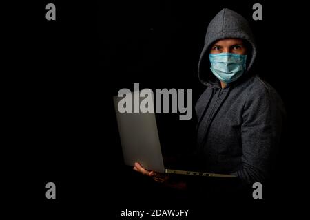 Man in a mask and a hood holds a laptop in his hands on a dark background. Concept of cyber rogue, hacker. Banner. Stock Photo