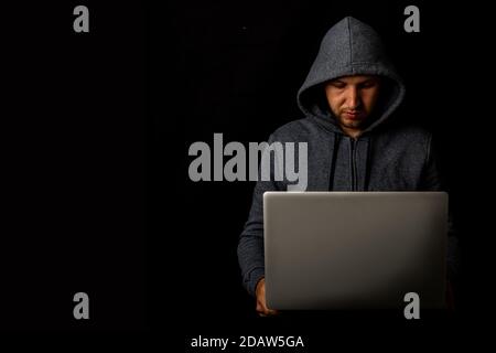 Man in a hood holds a laptop in his hands on a dark background. Concept of cyber rogue, hacker. Banner. Stock Photo