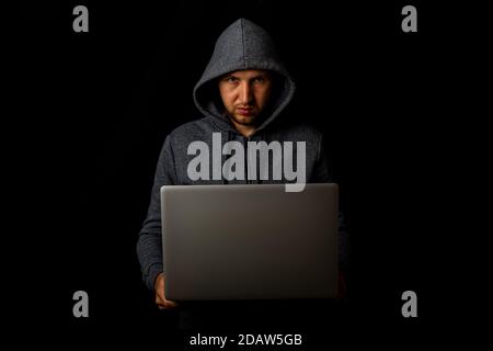 Man in a hood holds a laptop in his hands on a dark background. Concept of cyber rogue, hacker. Banner. Stock Photo