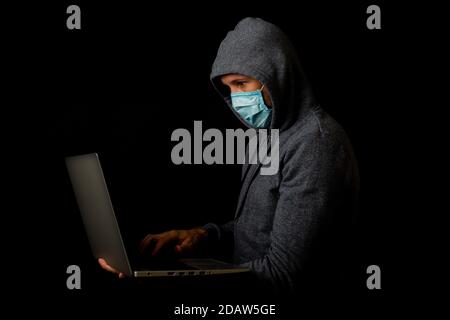 Man in a mask and a hood holds a laptop in his hands on a dark background. Concept of cyber rogue, hacker. Banner. Stock Photo