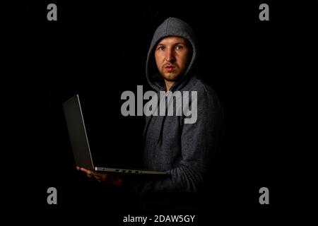 Man in a hood holds a laptop in his hands on a dark background. Concept of cyber rogue, hacker. Banner. Stock Photo