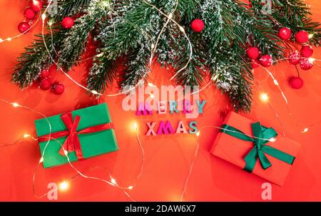 Xmas greeting card with fir branches, snow, berries, Christmas lights, gift boxes with color letters Merry Xmas on red. Stock Photo