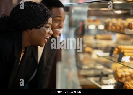 Surprised happy millennial woman and man standing in front of glass showcase with pastries and cakes Stock Photo