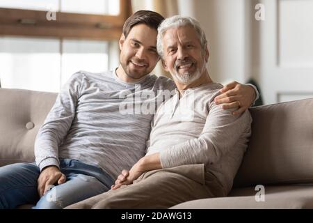 Portrait smiling young man hugging mature father, sitting on couch Stock Photo