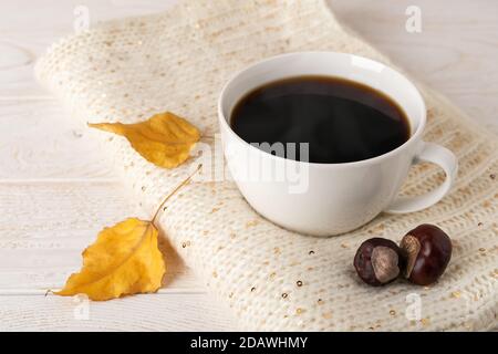 Large white mug of hot steaming coffee on a beige knitted scarf and two yellow fall leaves over white wood rustic surface. Cozy autumn mood concept. Stock Photo