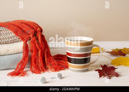Striped mug of hot steaming coffee or tea, stack of warm knitted sweaters and red fringed scarf and few yellow fall leaves over wood rustic surface. Stock Photo