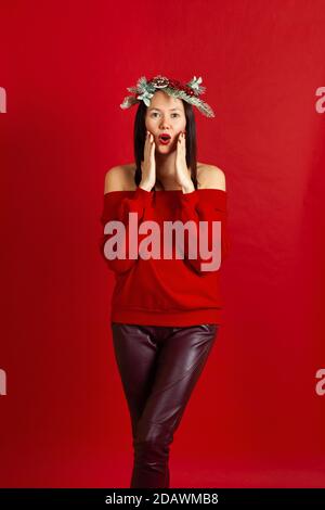full-length portrait of surprised Asian young woman with Christmas wreath with hands on her face on scarlet background Stock Photo