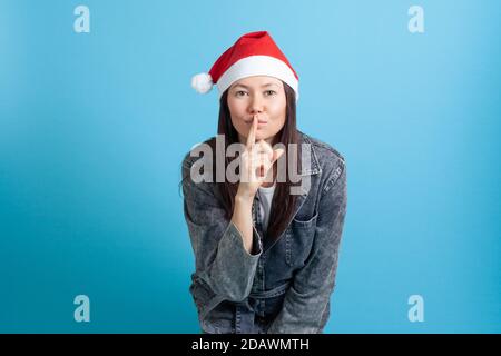 mock up portrait of a smiling Asian young woman in a Santa Claus hat puts her finger to her lips and asks for silence Stock Photo