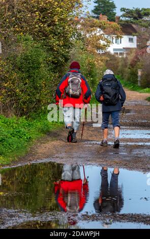 an older couple walking on a pathway through some puddles wearing winter waterproof clothing and rucksacks. Stock Photo