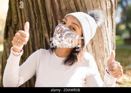 Portrait of young woman wearing a face mask. She has her thumbs up and is outdoors. Selective focus. Stock Photo